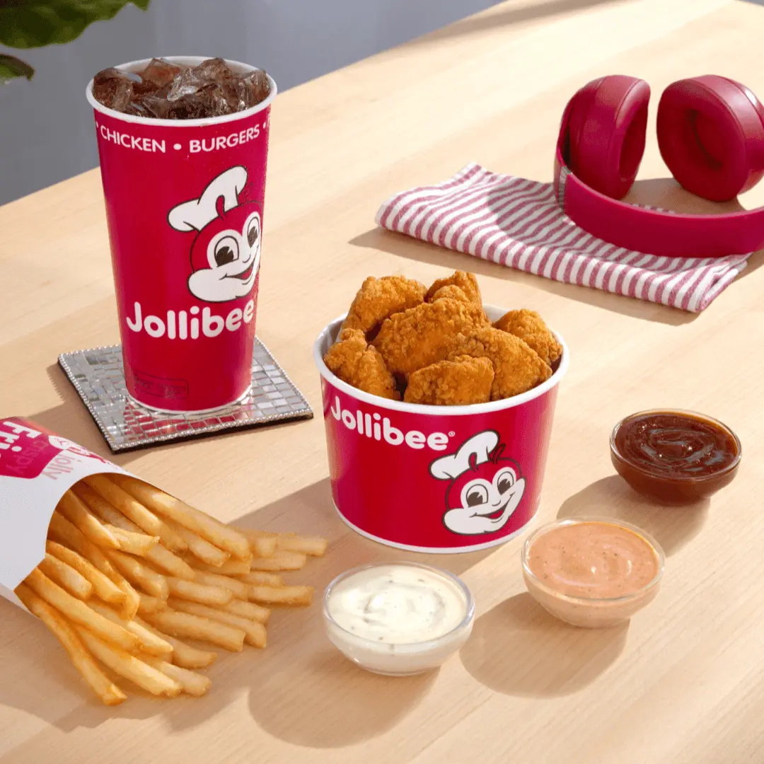 A red bucket of chicken nuggets with the Jollibee logo on it, with a drink in a red cup, a side of fries, and three different dips on a table.