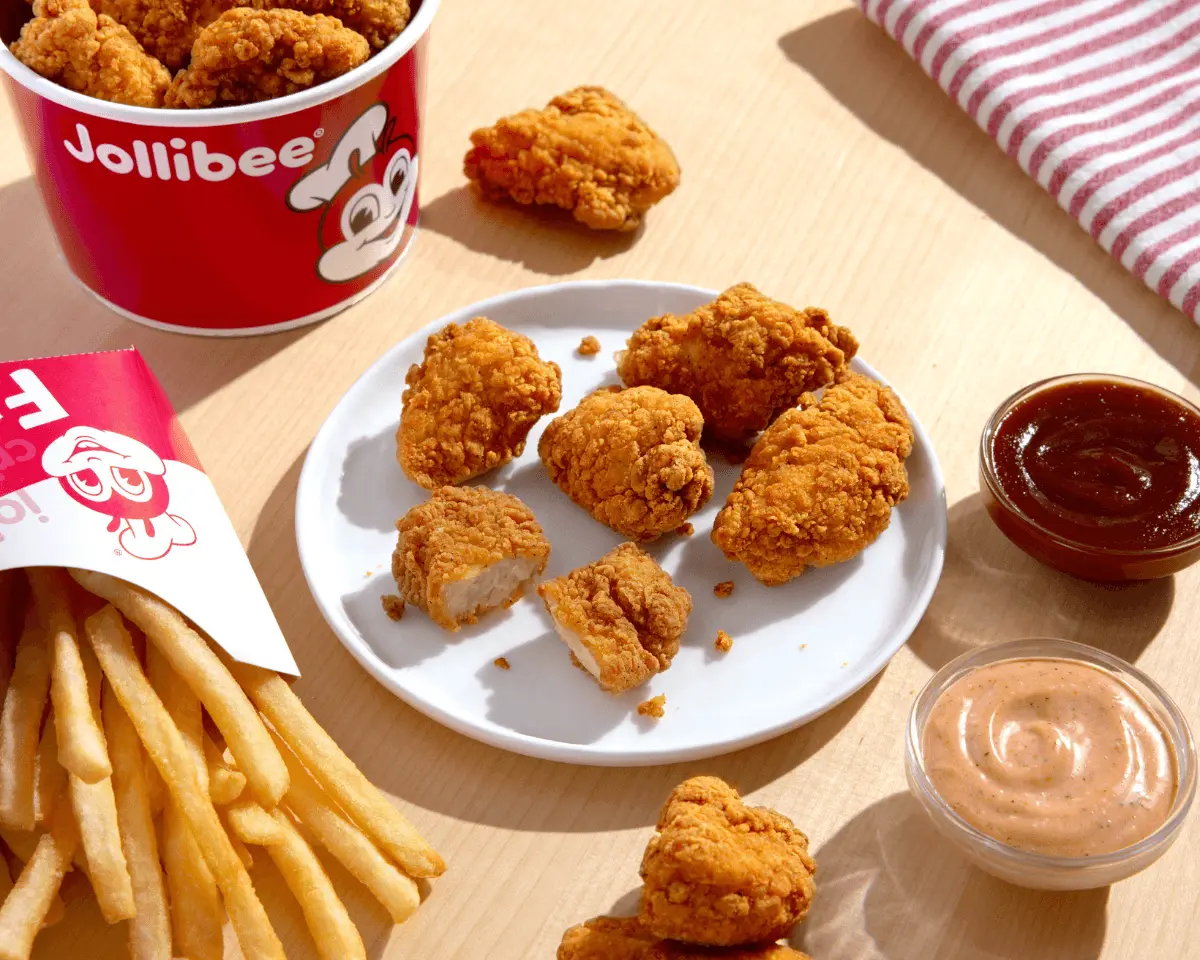 Chicken nuggets pieces on a white plate and some inside the red bucket with a side of fries and two dipping sauces on the table.